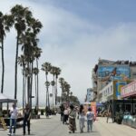 People walking along the Ocean Front Walk in Venice Beach with palm trees, shops, and restaurants on a cloudy day in Los Angeles.
