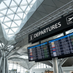 A low-angle photograph looking up at a large black digital departures sign in an airport terminal. The sign displays the word "DEPARTURES" in white next to an "i" icon and an airplane takeoff icon. Below it are smaller screens with flight information (Cid., To, Flight, Gate, Remarks) for destinations like MILANO, NEW YORK, and SYDNEY. The background shows a complex architectural ceiling structure with geometric metal and glass panels, supported by metal beam columns.